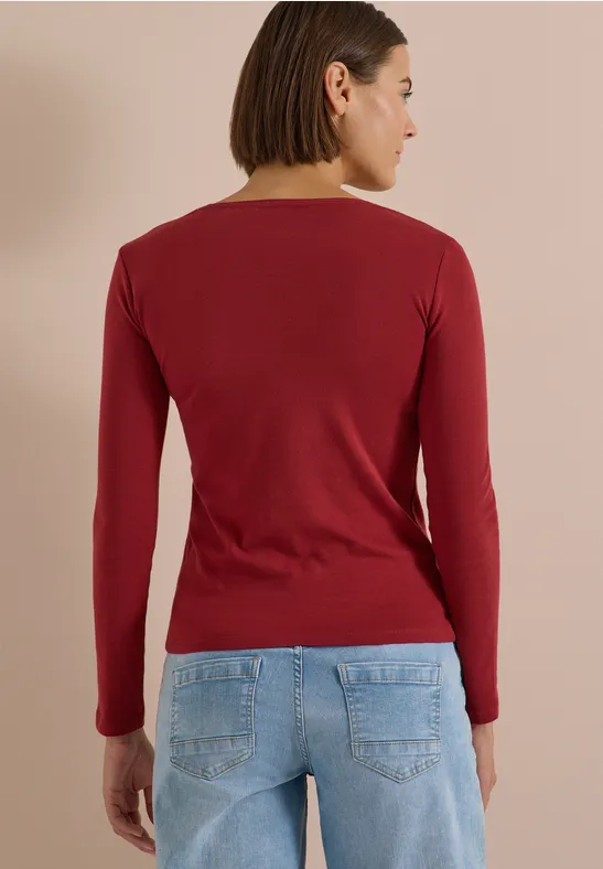 Femme vue de dos portant un tee-shirt à manches longues rouge et un jean bleu clair sur fond neutre.