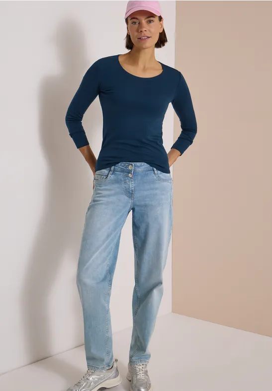 Femme en jean clair, haut bleu et casquette rose, appuyée de manière décontractée contre un mur.