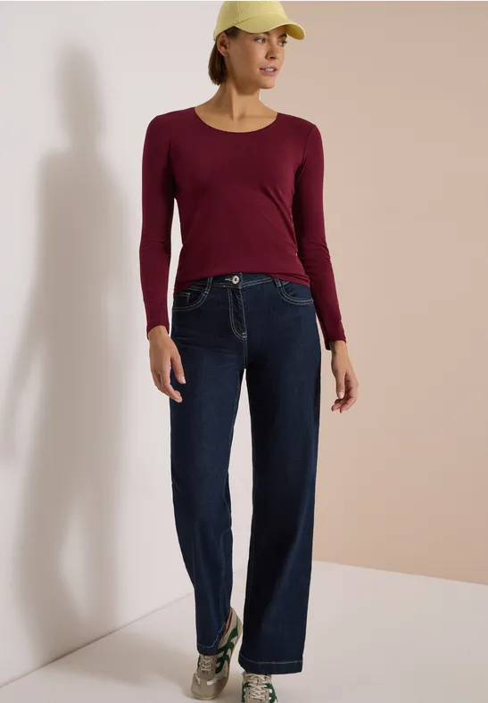 Femme portant un haut bordeaux, un jean bleu foncé large, des baskets et une casquette, appuyée sur un mur beige.