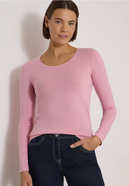 Femme en haut rose à manches longues et jean foncé, souriante, main dans la poche.