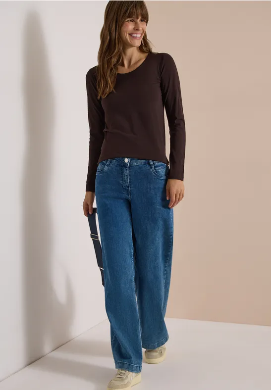 Frau in braunem Langarmshirt, weiten blauen Jeans und hellen Sneakers lächelt vor neutralem Hintergrund.