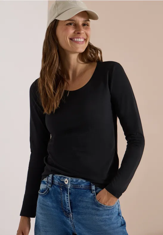 Eine lächelnde Frau in schwarzem Langarmshirt, blauen Jeans und beiger Basecap, Hand in Tasche.