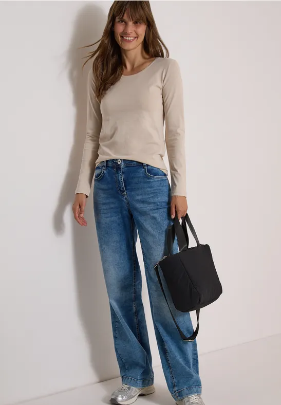 Frau trägt Jeans, beiges Langarmshirt, schwarze Tasche, lächelt vor neutralem Hintergrund.