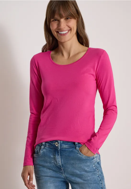 Lächelnde Frau in pinkem Langarmshirt und Jeans vor neutralem Hintergrund.