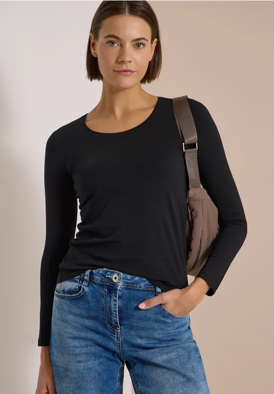 Femme portant un top noir à manches longues, un jean bleu et un sac épaule sur fond neutre.
