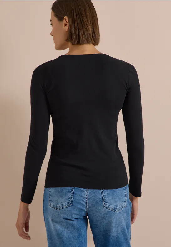 Une femme de dos porte un t-shirt à manches longues noir et un jean bleu clair devant un fond beige.