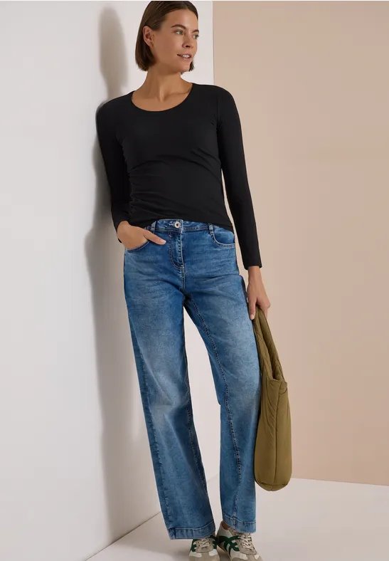 Femme appuyée contre un mur blanc, porte un haut noir, un jean droit bleu et tient un sac vert.