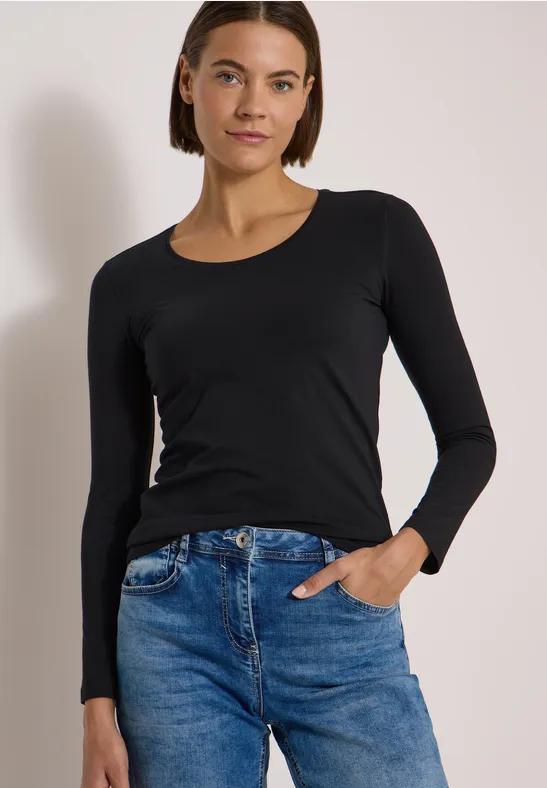 Femme portant un t-shirt noir à manches longues et un jean bleu, posant décontractée devant un fond clair.