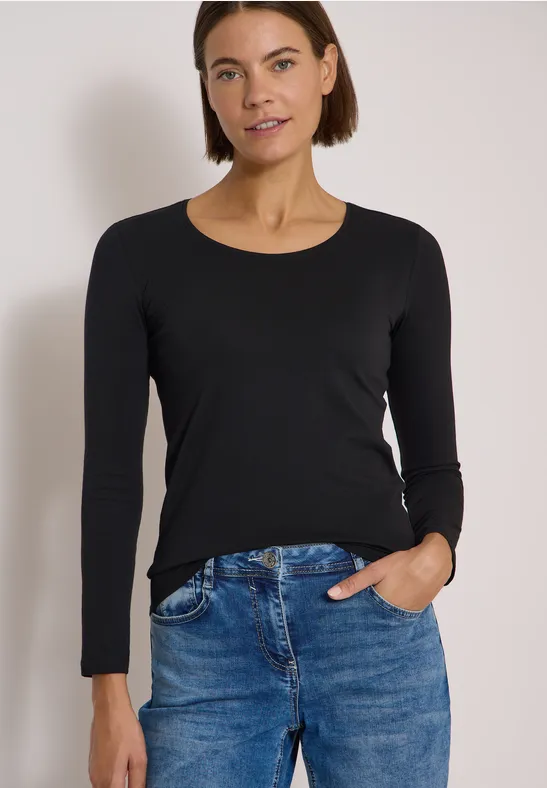 Une femme en t-shirt noir à manches longues et jean bleu pose décontractée, main dans la poche, devant un mur clair.