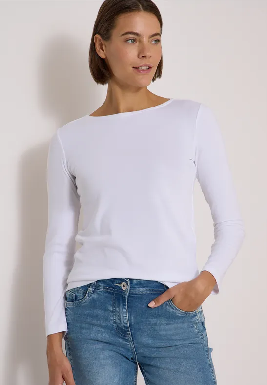 Femme portant un haut blanc à manches longues et un jean bleu, une main dans la poche, regard de profil.