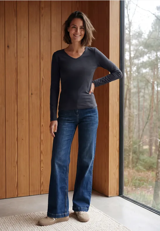 Eine lächelnde Frau in langärmeligem Shirt und weiten Jeans steht vor einer Holz-Wand und Fenster mit Waldblick.