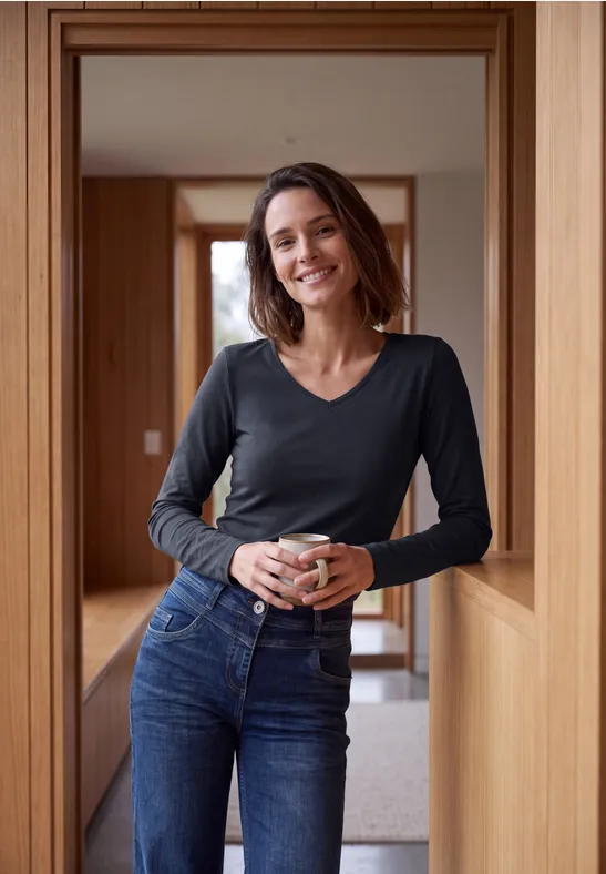 Lächelnde Frau in Jeans und Langarmshirt lehnt an Holzrahmen und hält eine Tasse.