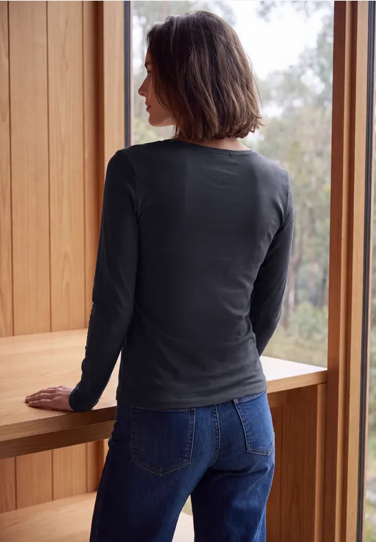 Eine Frau mit dunklen Haaren steht an einem Fenster in einem Holzrahmen, trägt Jeans und langärmeliges Shirt.