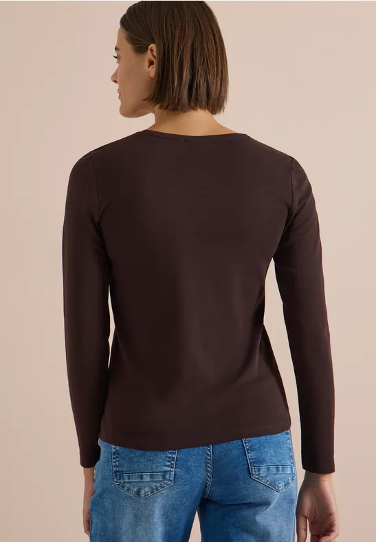 Eine Frau steht von hinten, trägt einen dunkelbraunen Langarmpullover und blaue Jeans.