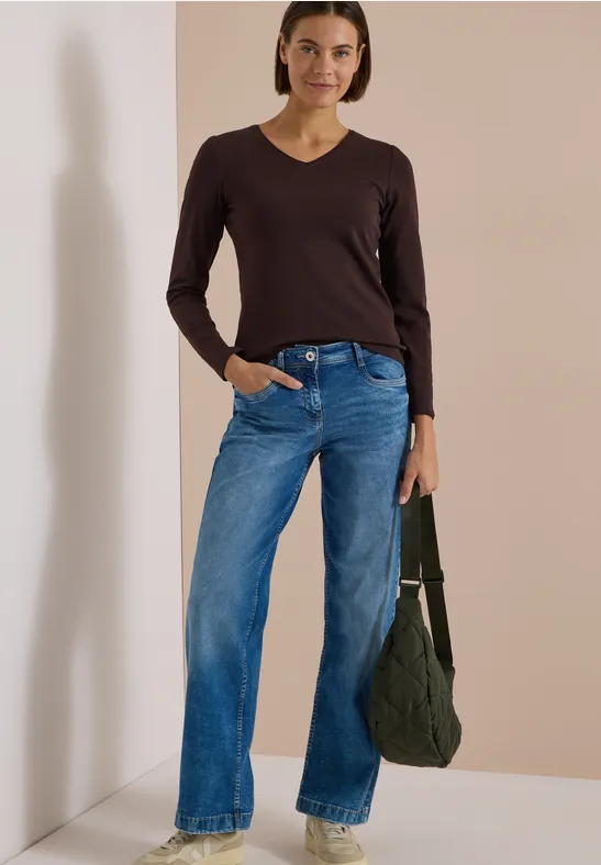 Frau in braunem V-Ausschnitt-Pullover, weiten blauen Jeans und grüner Tragetasche steht vor beigem Hintergrund.