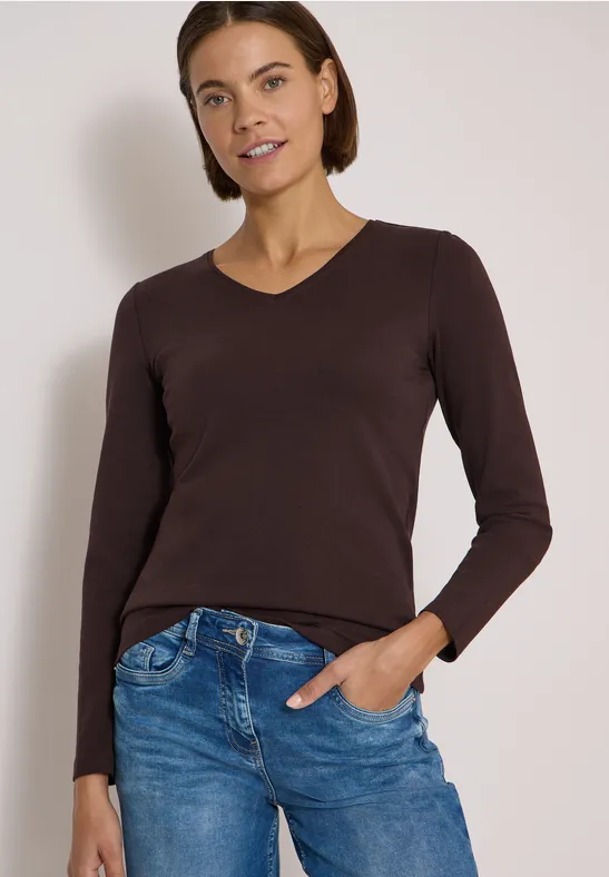 Junge Frau mit braunem V-Ausschnitt-Langarmshirt und blauer Jeans, lächelt, Hand in der Tasche.