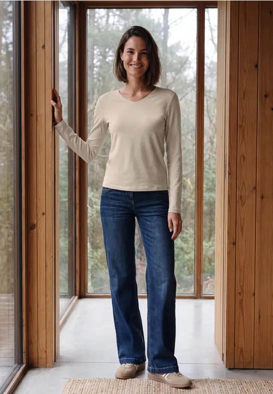 Eine lächelnde Frau in hellem Pullover und Jeans steht vor bodentiefen Holzfenstern.