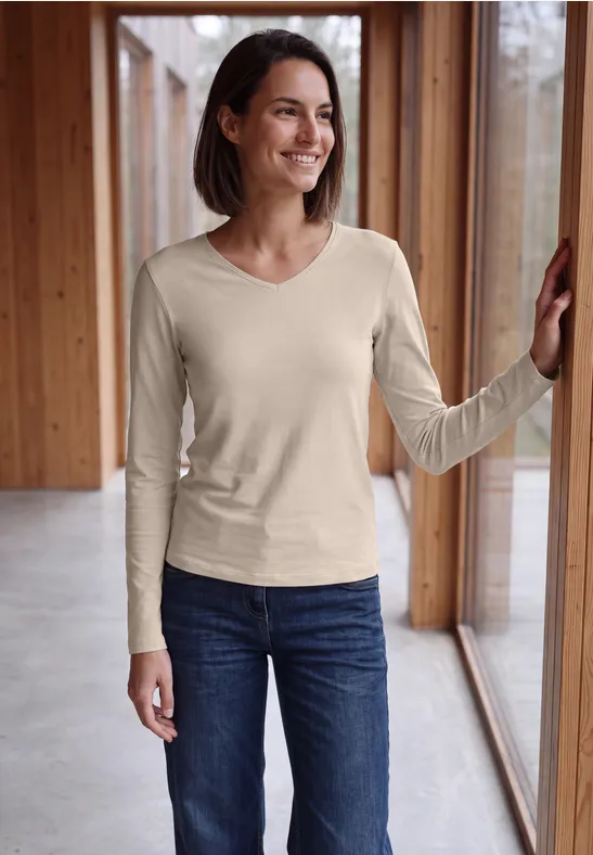 Lächelnde Frau im beigen Langarmshirt und Jeans lehnt an Fensterrahmen in modernem Holzinterieur.