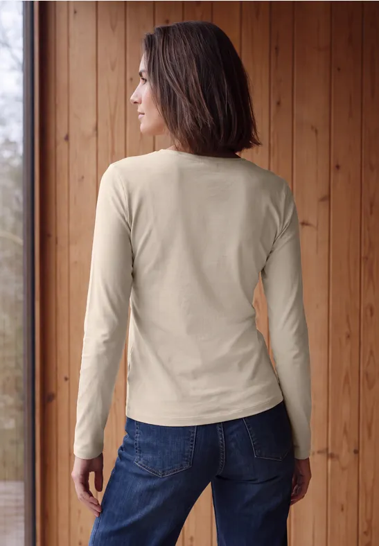 Frau in langärmeligem beigem Shirt und Jeans, Rückansicht vor einer Holzverkleidung.