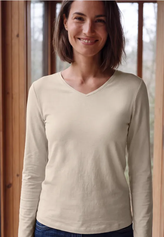 Eine lächelnde Frau vor einem Fenster, trägt cremefarbenes, langärmliges V-Neck-Shirt.