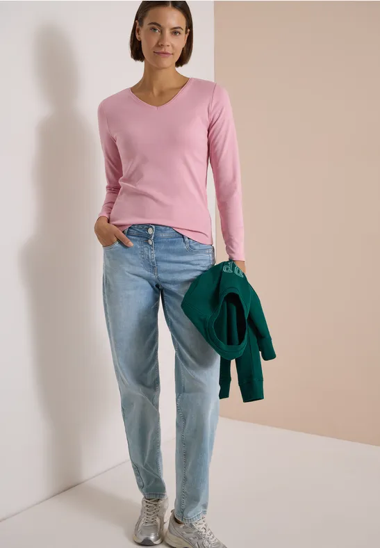 Frau steht vor neutralem Hintergrund in rosa Pullover, Jeans und grauen Sneakern, hält grüne Jacke.