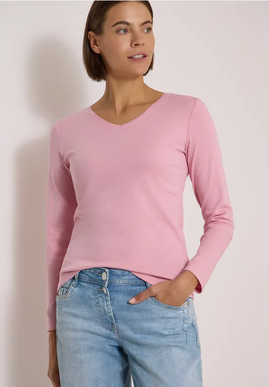 Frau trägt pinken V-Ausschnitt-Pullover und Jeans, steht vor heller Wand.