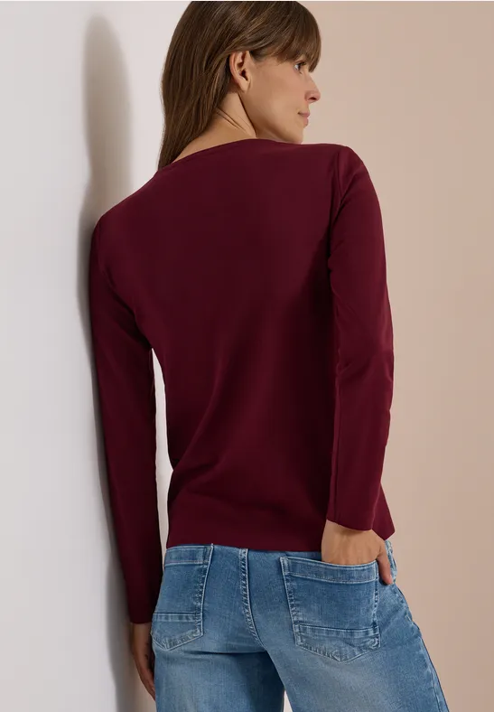 Frau von hinten in bordeauxrotem Pullover und hellblauen Jeans, mit einer Hand in der Gesäßtasche.