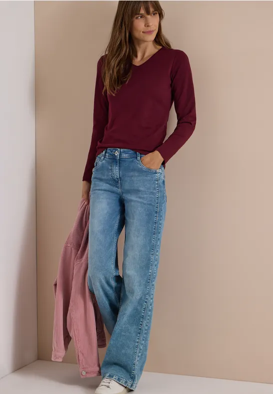 Frau in weiten Jeans und burgunderfarbenem Longsleeve, hält rosa Jacke, vor schlichtem beigem Hintergrund.