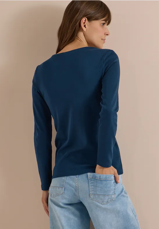 Frau von hinten in dunkelblauem Langarmshirt und hellen Jeans vor beigem Hintergrund.