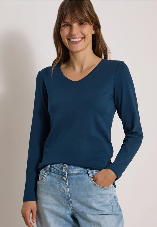 Frau mit blauem Langarmshirt und Jeans lächelt vor hellem Hintergrund.