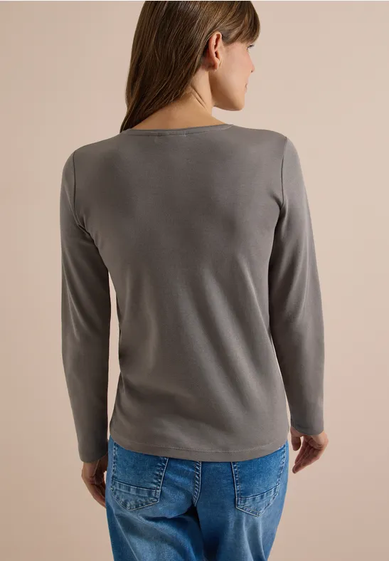 Frau von hinten in grauem Langarmshirt und blauen Jeans vor neutralem Hintergrund.
