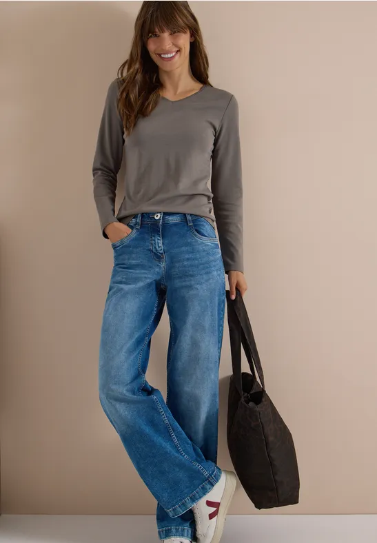 Modisch gekleidete Frau in langärmeligem Shirt, weiten Jeans und Tasche, lächelt vor neutralem Hintergrund.