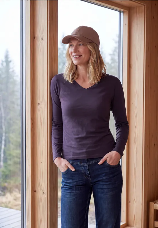 Lächelnde Frau mit Cap, dunkelblauem Shirt und Jeans steht in Holzhütte vor großem Fenster mit Waldblick.