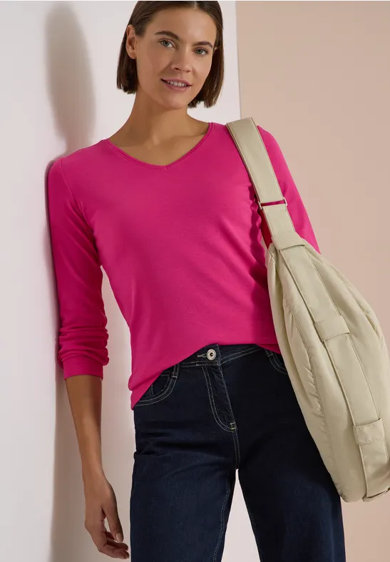 Frau lehnt vor hellem Hintergrund, trägt pinkes Longsleeve, dunkle Jeans und beige Schultertasche.