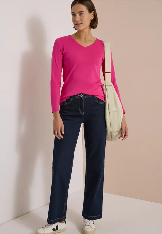Frau mit pinkem Pullover, weiten Jeans und beiger Tasche vor hellem Hintergrund.