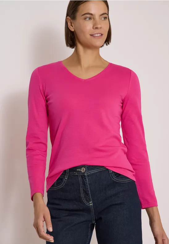 Frau in pinkem V-Ausschnitt-Pullover und dunklen Jeans, die lächelnd in die Ferne schaut.