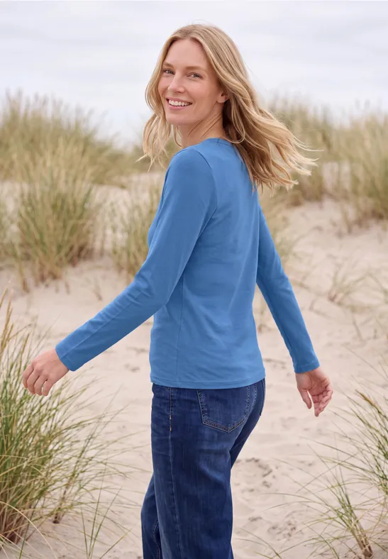 Eine lächelnde Frau in blauem Pullover und Jeans steht in Dünen mit Strandgras.