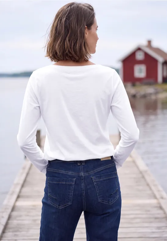 Person mit weißem Langarmshirt und Jeans steht auf Holzsteg, Blick aufs Wasser und rotes Haus.
