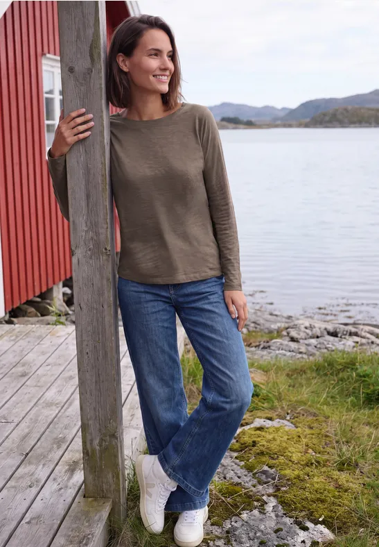 Frau lehnt lächelnd an einem Holzpfosten am Seeufer, trägt Pullover und Jeans.