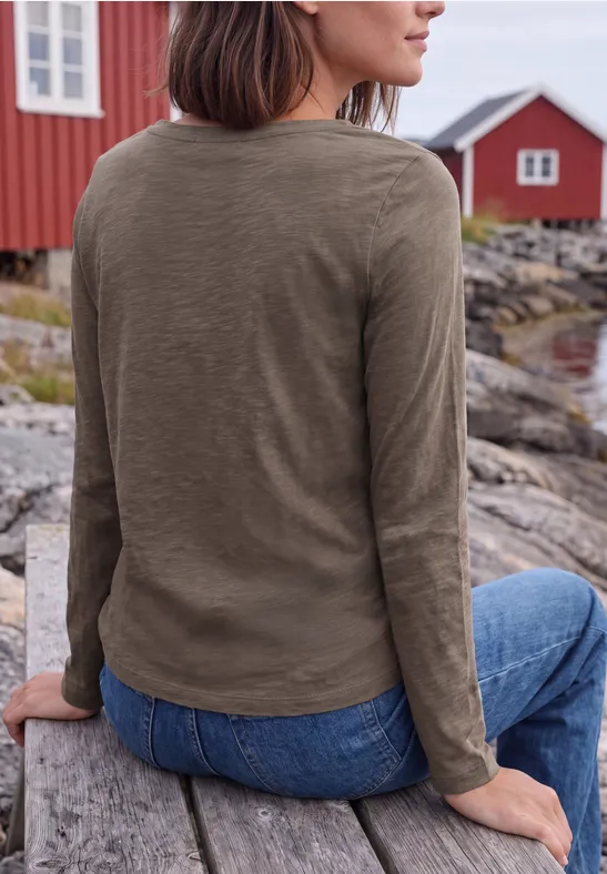 Frau sitzt auf Holzbank am Wasser, trägt olivgrünes Shirt und Jeans, rote Häuser im Hintergrund.