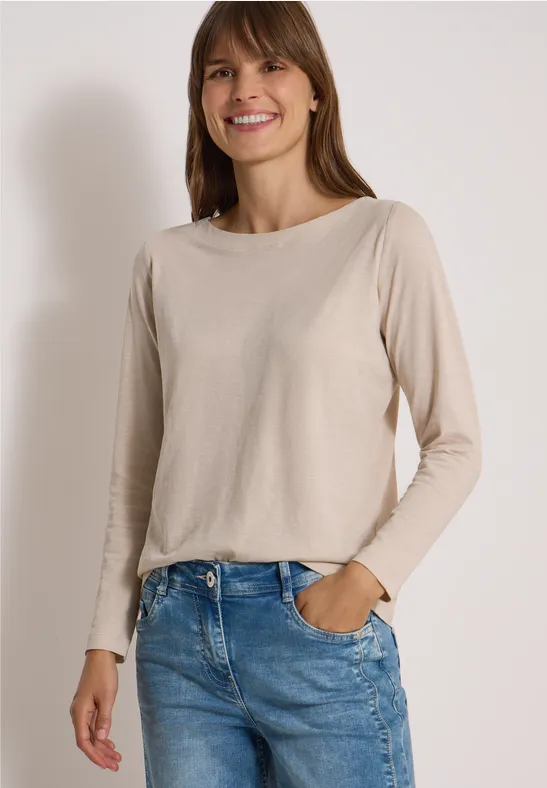 Lächelnde Frau in beige langärmligem Shirt und blauen Jeans, steht mit einer Hand in der Tasche.