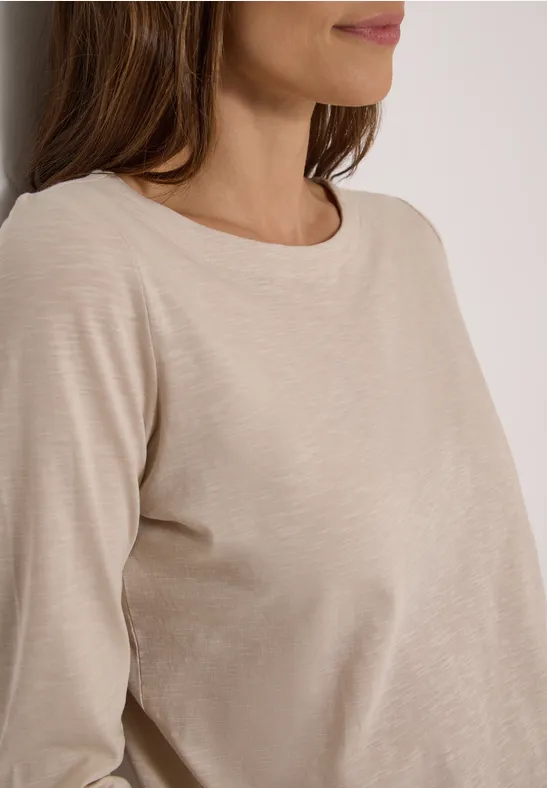Frau mit beigem Langarmshirt lehnt sich an eine weiße Wand.