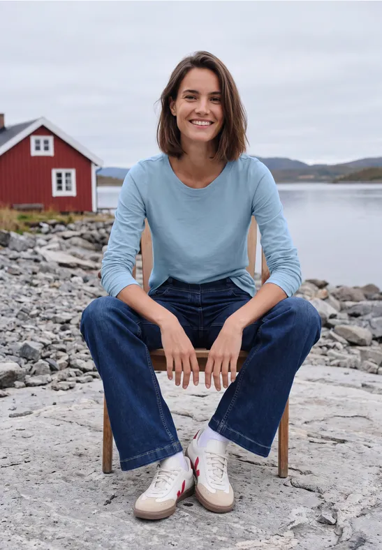 Eine lächelnde Frau in Jeans und blauem Shirt sitzt auf einem Stuhl am felsigen Ufer mit rotem Haus im Hintergrund.