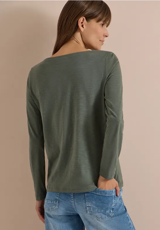 Frau mit langen braunen Haaren in olivgrünem Langarmshirt, Rückenansicht, Hände in Jeans-Taschen.