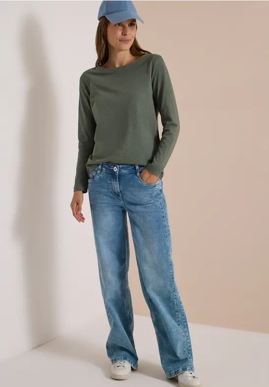 Frau in weitem Jeans, grünem Langarmshirt, weißen Sneakern und blauer Baseballkappe vor hellem Hintergrund.