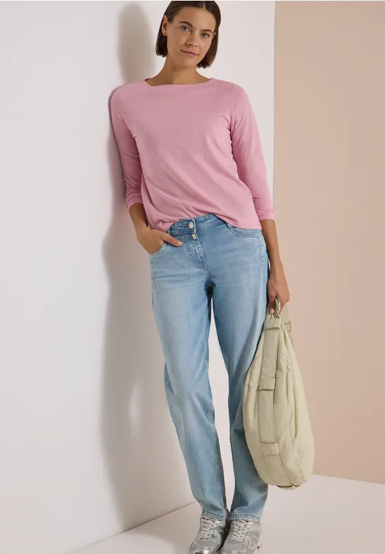 Frau in pinkem Shirt und blauen Jeans lehnt lächelnd an der Wand und hält eine große Tasche.