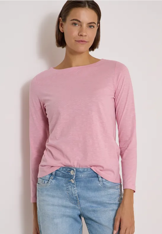 Frau in rosa Langarmshirt und blauen Jeans vor hellem Hintergrund.