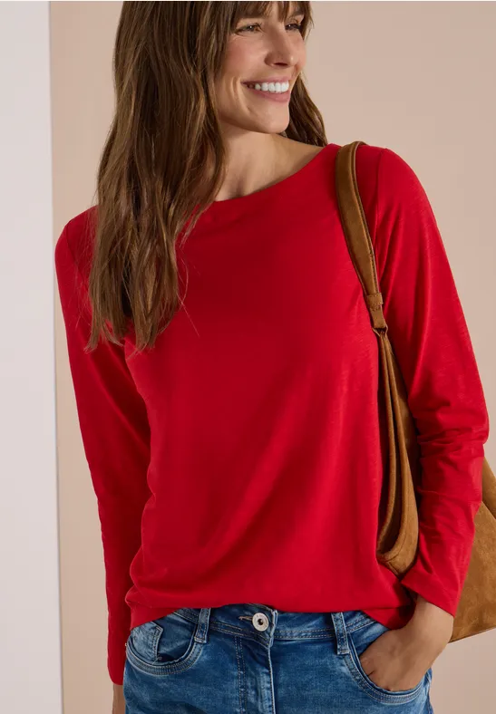 Lächelnde Frau im roten Langarmshirt trägt lockere Hose und Schultertasche vor neutralem Hintergrund.