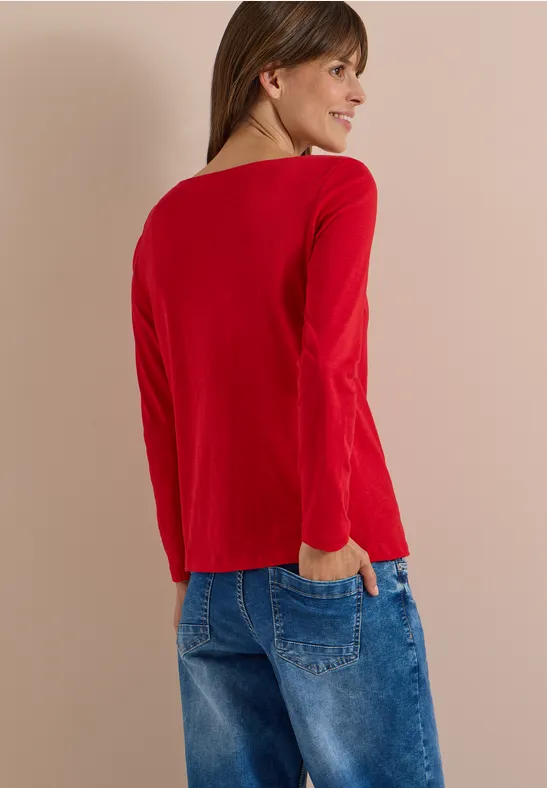 Eine Frau steht von hinten in einem roten Langarmshirt und blauen Jeans vor hellem Hintergrund.