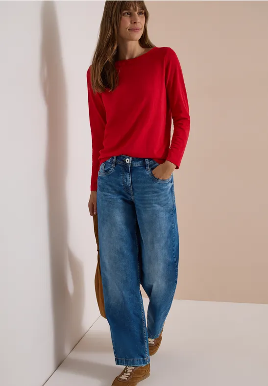 Frau in rotem Pullover und weiter Jeanshose vor neutralem Hintergrund.
